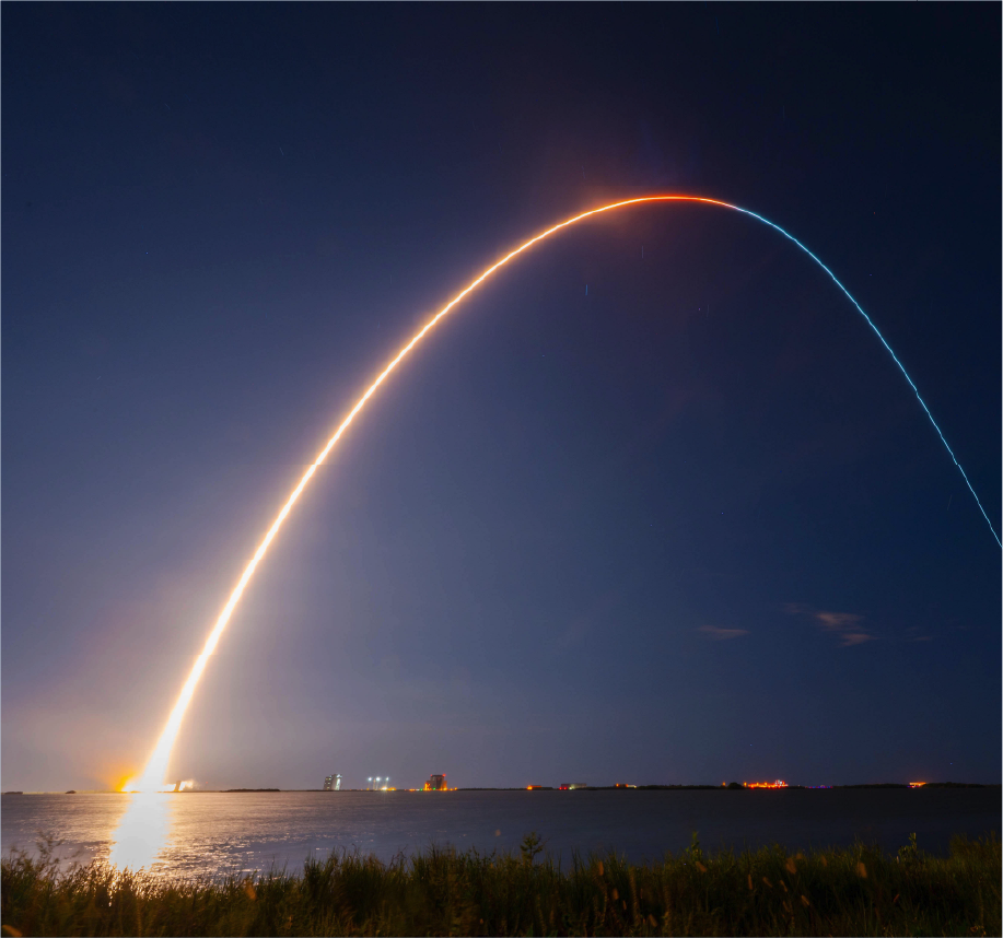 A bright arc of light streaks across a night sky, showing a rocket launch over water—a powerful visual of technology and its applications—with grassy shoreline in the foreground and distant lights on the horizon.